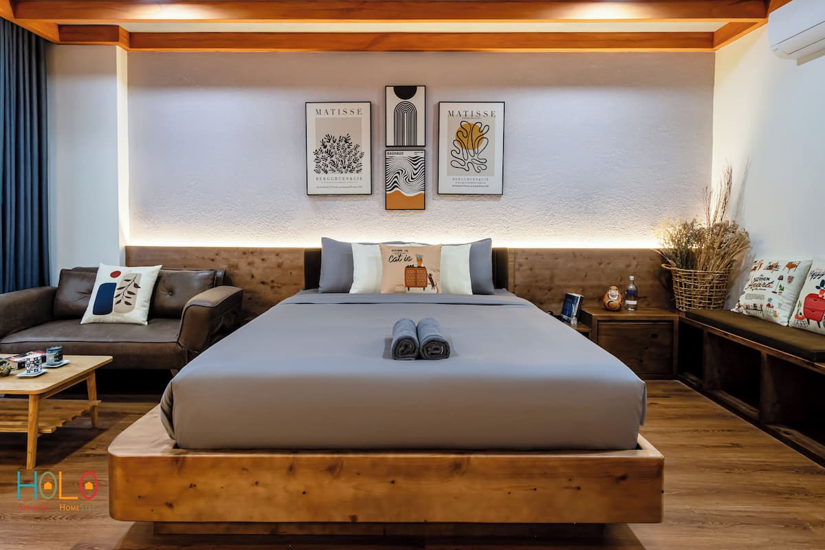 A modern bedroom with a large bed, a sofa, and a small table. There are wall-mounted artworks and a basket of dried plants.