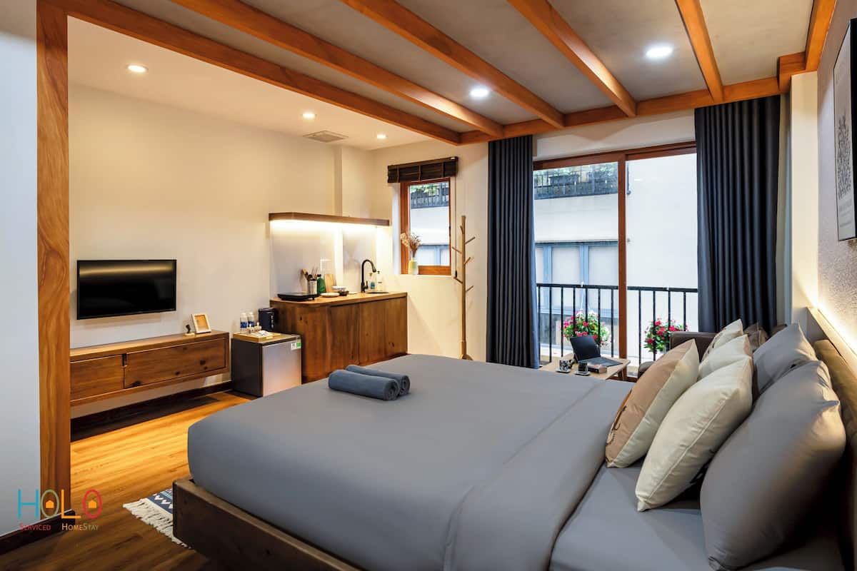 A modern hotel room with a large bed, wooden beams, a TV, a desk, and a balcony with flowers.