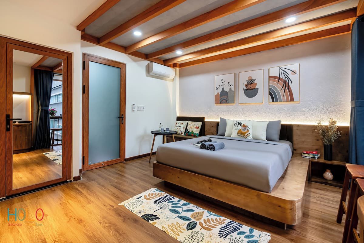 A modern bedroom with a wooden bed frame, a small bedside table, and a nightstand. There is a window with curtains, a wall-mounted air conditioner, and two framed artworks above the bed.