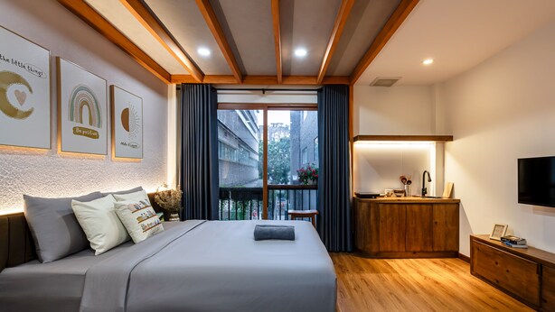 A modern bedroom with a large bed, a wooden headboard, a bedside table with a lamp, a TV, and a view of the city through a balcony.