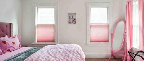 A bedroom with a bed, a dresser, a mirror, and two windows with pink curtains.