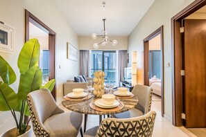 A dining area with a round table set for four, surrounded by chairs. A large plant is visible to the left, and a modern chandelier hangs from the ceiling. - Enticing 2BR Apartment in Reva Residences, Business Bay by Deluxe Holiday Homes (Dubai)