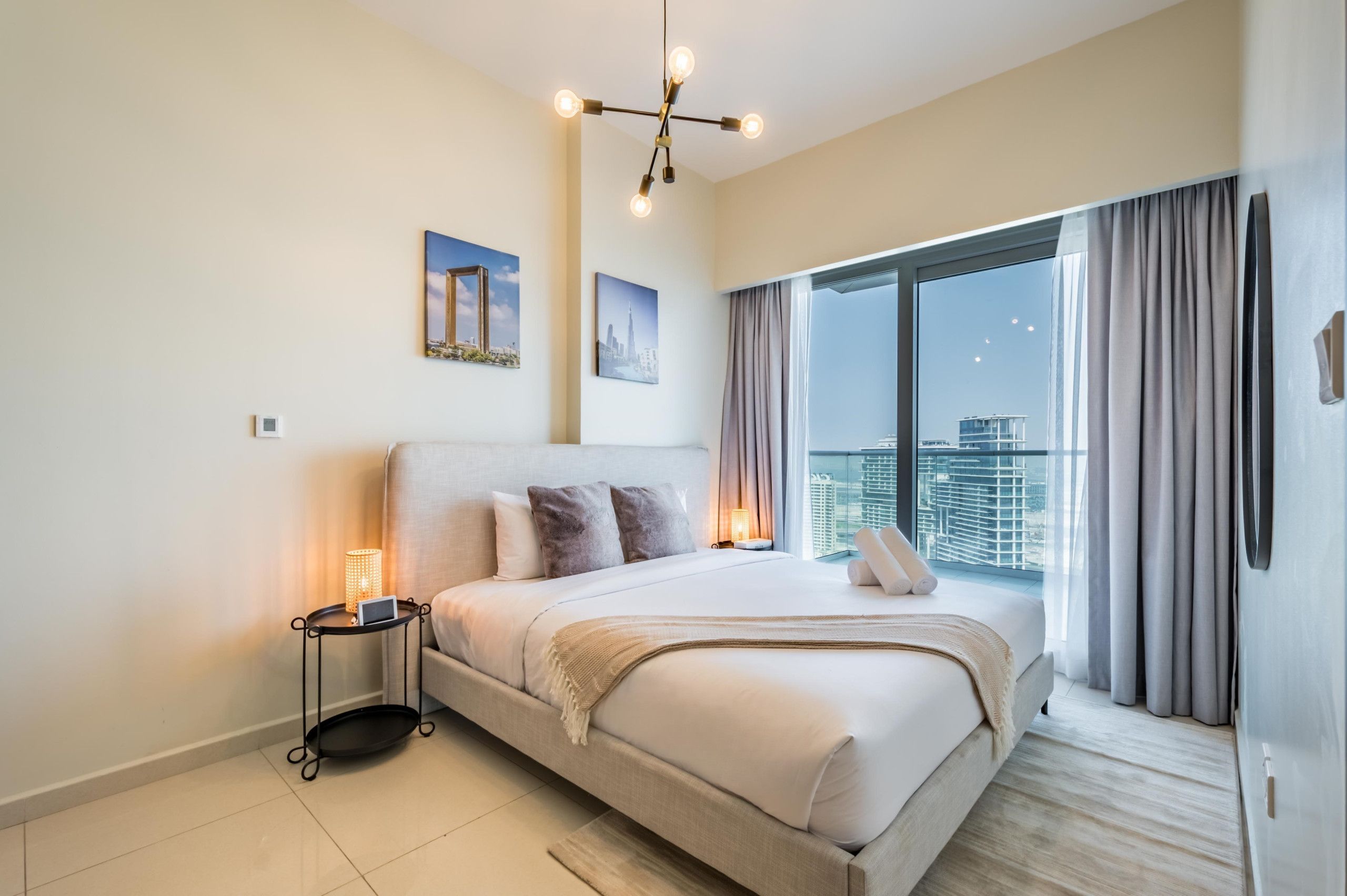 Une chambre moderne avec un grand lit, une table de chevet avec une lampe et une vue sur la ville à travers de grandes fenêtres.