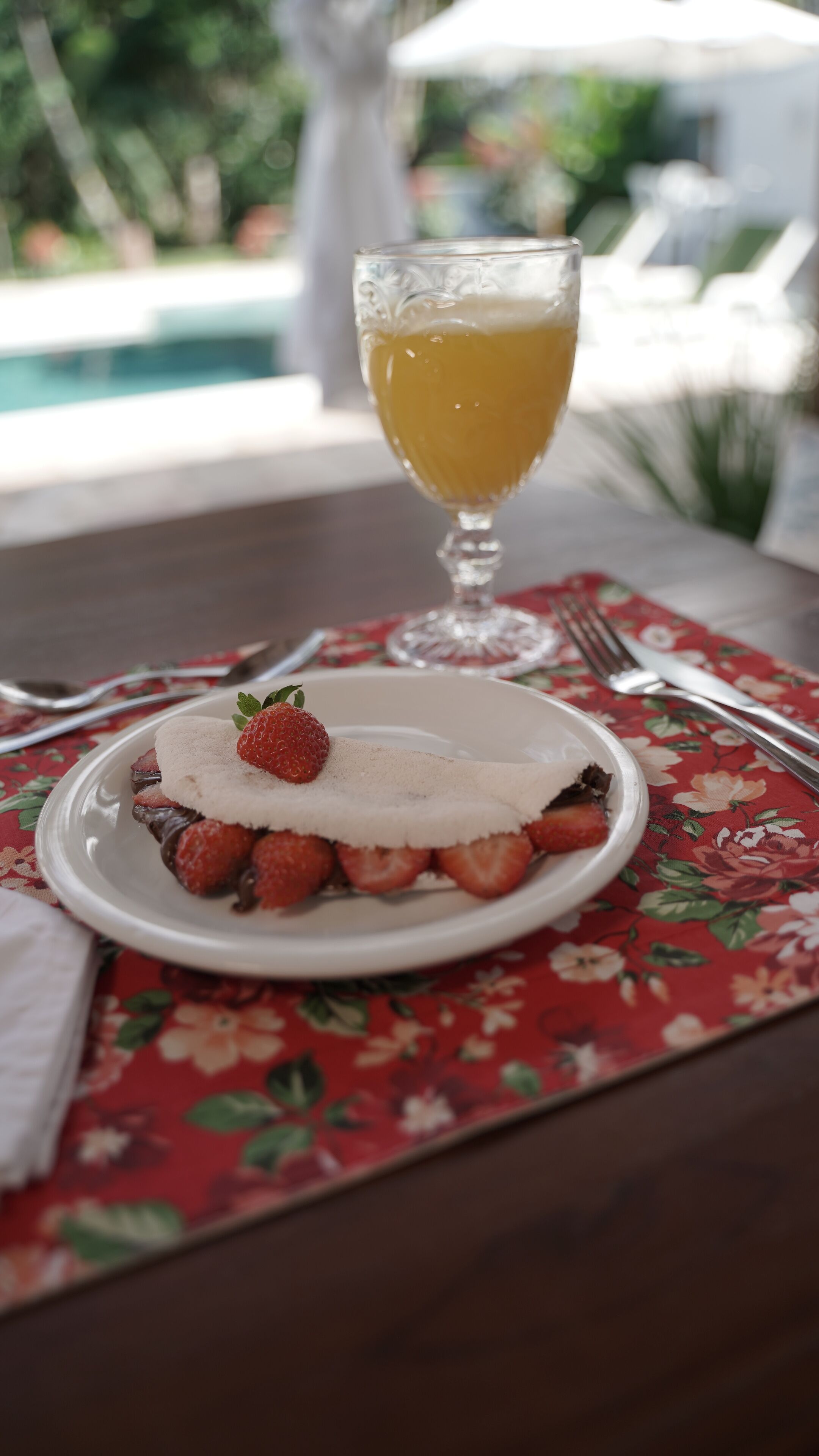 A plate with a dessert topped with strawberries and whipped cream, a glass of orange juice, and a fork and knife on a table with a floral tablecloth.