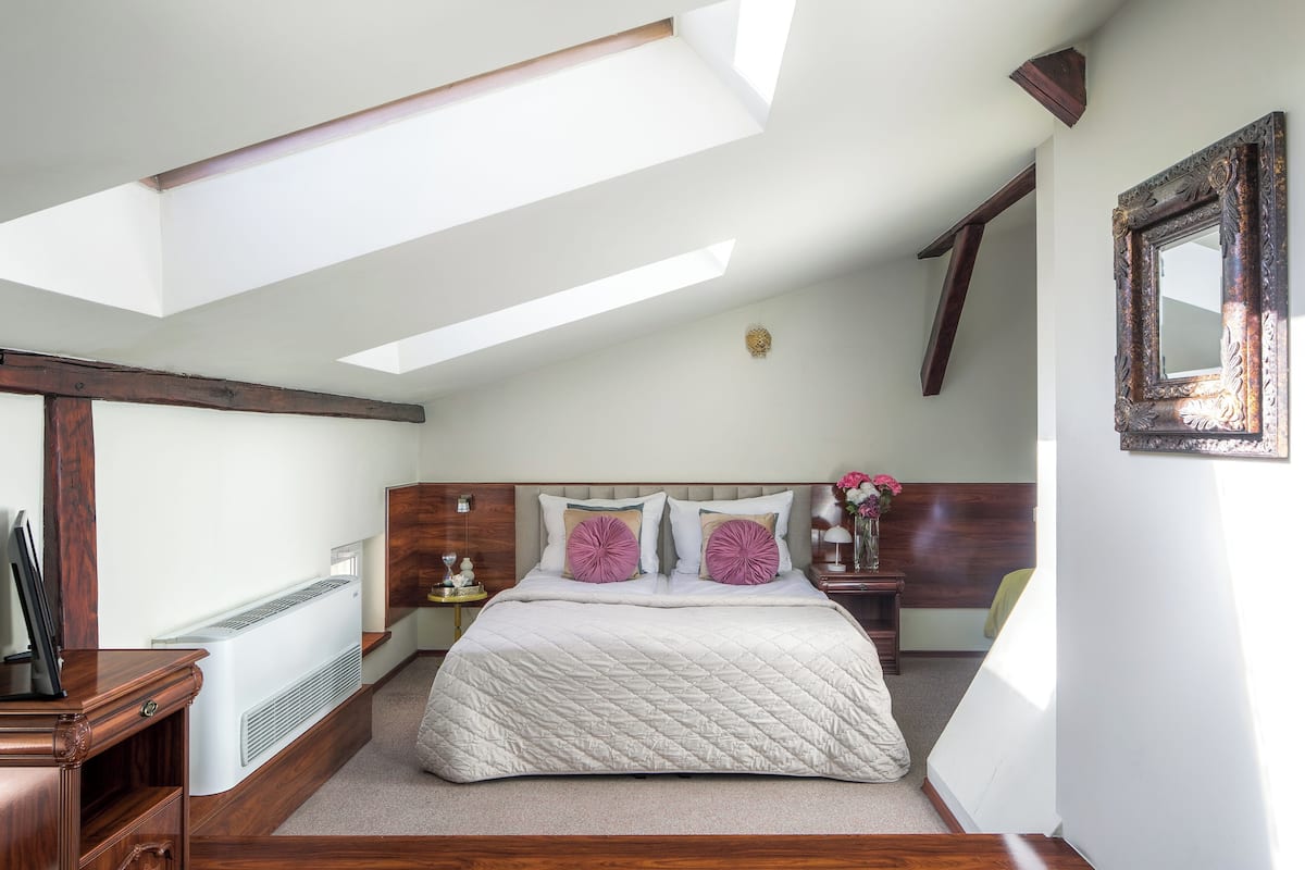 A bedroom with a sloped ceiling, a bed with a quilted duvet, a bedside table with a lamp, and a wall-mounted mirror.