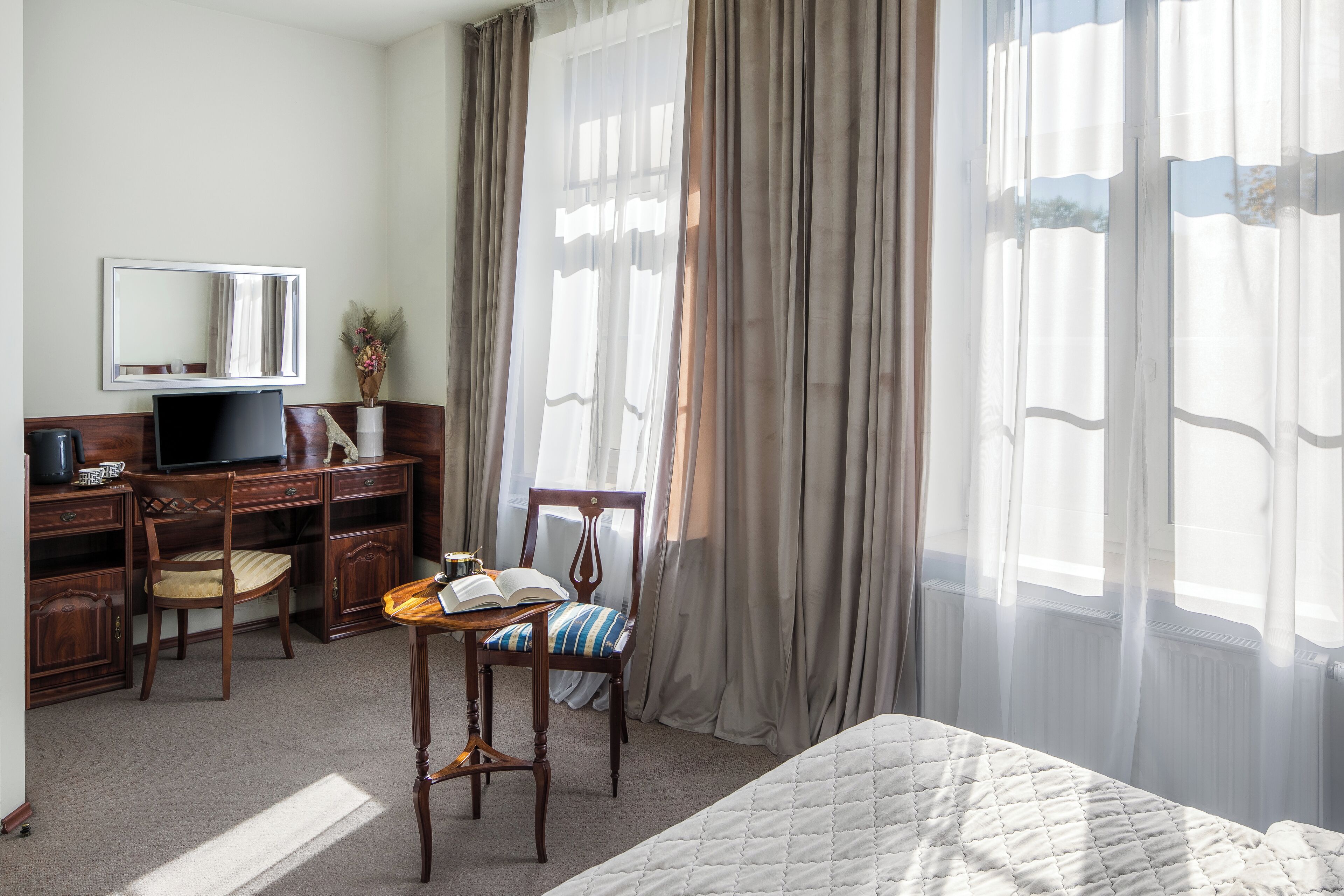 Habitación de hotel con cama, escritorio con computadora, silla, mesita con un libro, espejo y cortinas traslúcidas.