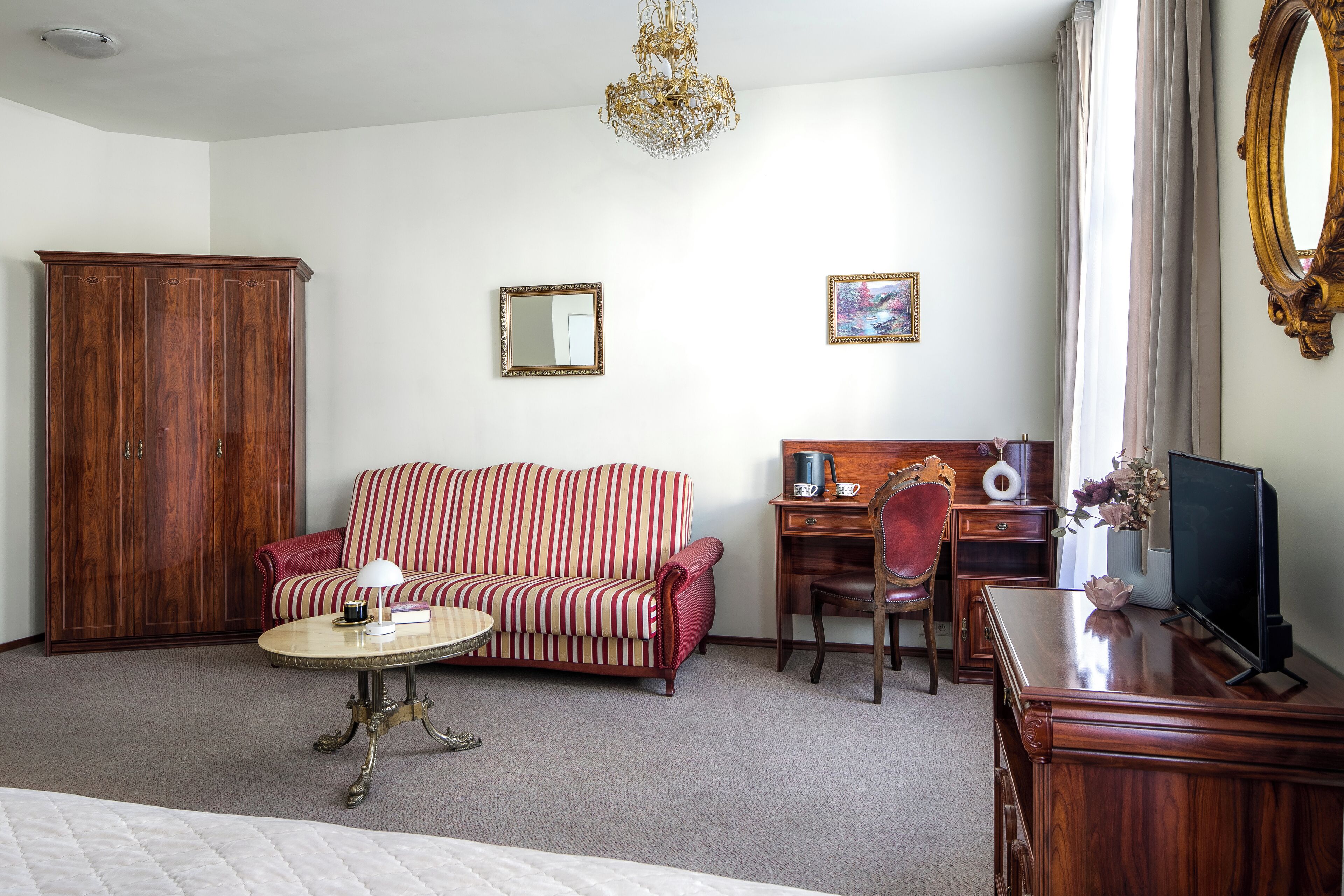 Habitación de hotel con un armario de madera, un sofá a rayas, una mesa de centro redonda, un escritorio con una silla, un televisor y una araña.