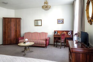 Habitación de hotel con un armario de madera, un sofá a rayas, una mesa de centro redonda, un escritorio con una silla, un televisor y una araña.