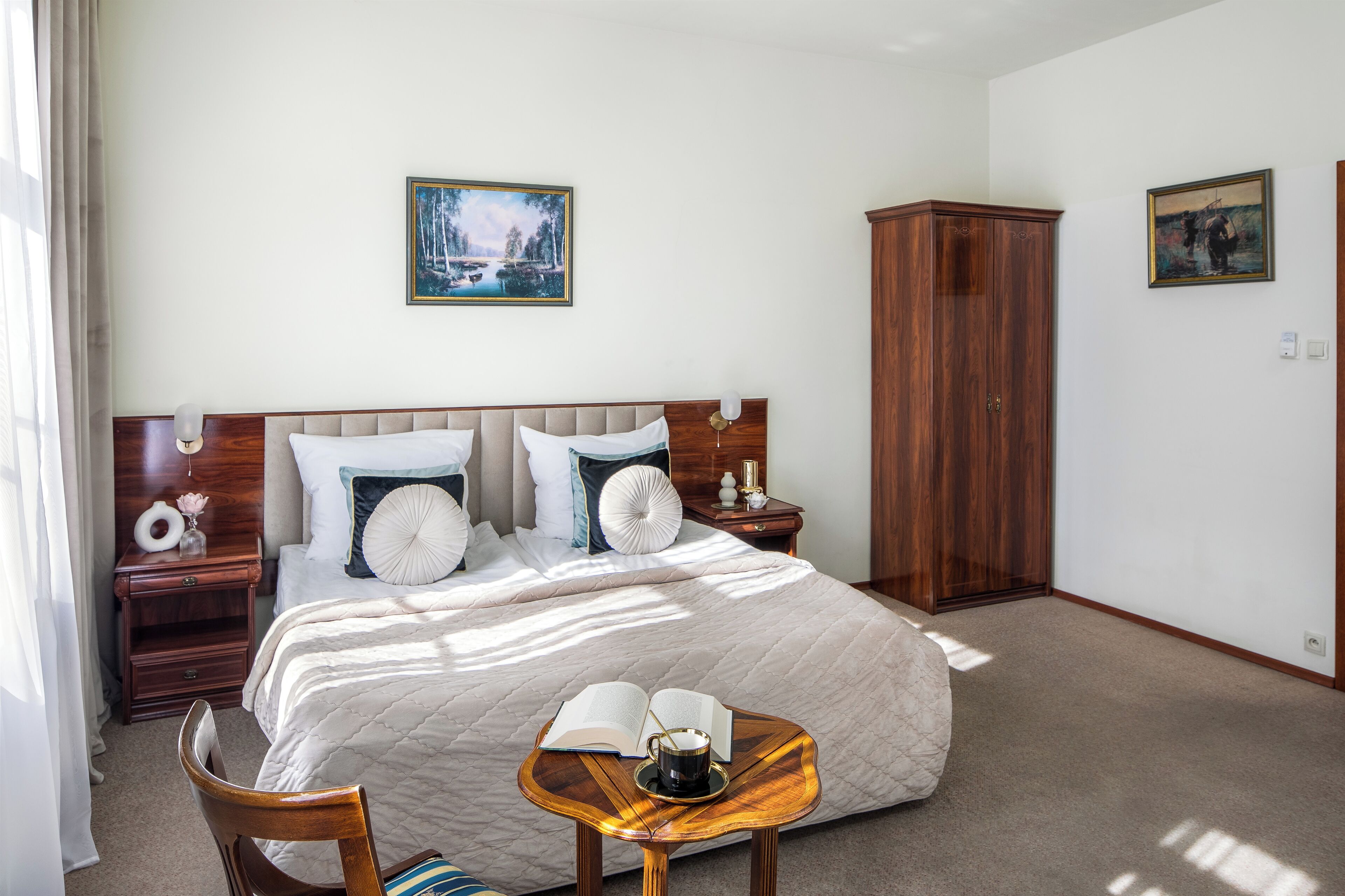 Une chambre à coucher bien rangée, avec un grand lit, une table de chevet avec une lampe, une petite table en bois sur laquelle se trouvent un livre et une tasse, et deux tableaux accrochés au mur.