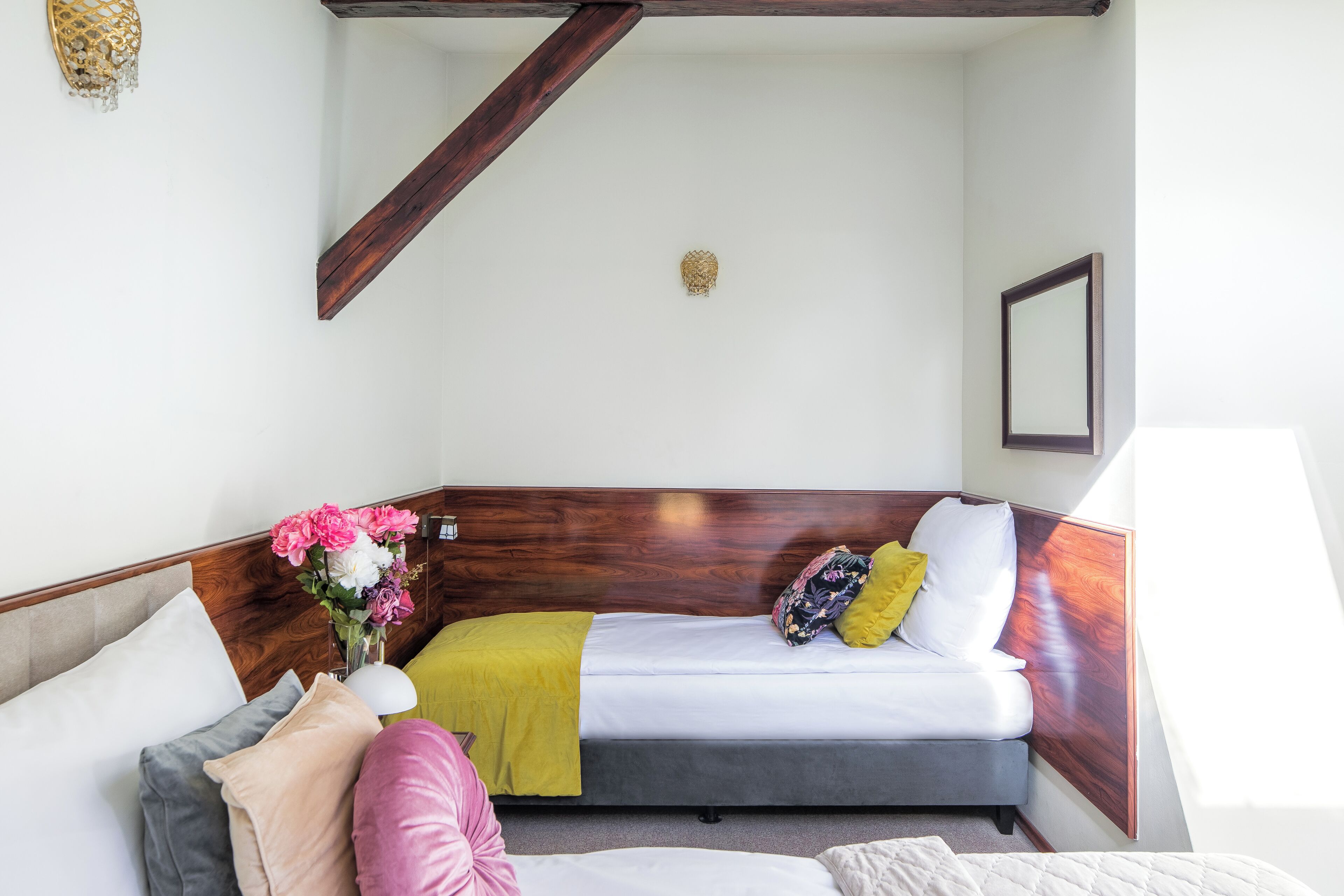A hotel room with a bed, a bedside table with a lamp, a wooden headboard, and a wall-mounted light fixture.