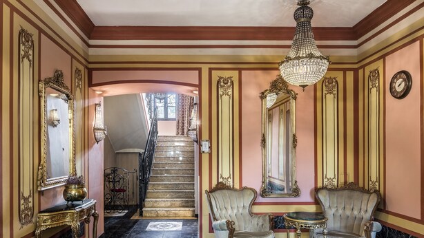 Un amplio pasillo con espejos dorados ornamentados, un candelabro y una escalera que conduce a un nivel superior.