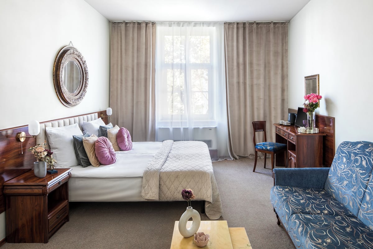 A bedroom with a bed, a desk, a chair, a sofa, a mirror, and a window with curtains.