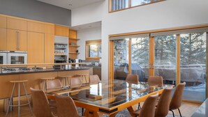 A modern kitchen with wooden cabinets, a glass dining table, and a view of a snowy landscape through large windows. - Gray Mill House-Curate Telluride-New Remodel, Hot Tub, Grill, Views, Lux Home (Mountain Village)