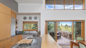 A modern kitchen with a central island, wooden cabinets, and a large window overlooking a patio area. - Gray Mill House-Curate Telluride-New Remodel, Hot Tub, Grill, Views, Lux Home (Mountain Village)