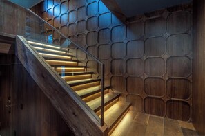 Una escalera moderna con escalones de madera y barandilla de vidrio, que lleva a un espacio con un diseño geométrico en la pared.