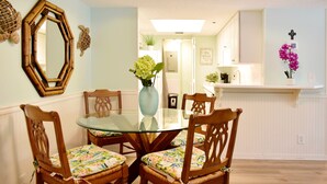 A dining area with a glass-top table surrounded by wooden chairs with floral cushions, a vase with flowers, and a wall-mounted mirror. - Beach Club 203 aka "Beach Walk" 2br/2ba Oceanfront Complex (Saint Simons Island)