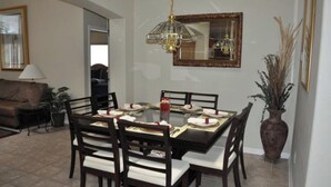 Un coin repas avec une table ronde dressée pour quatre personnes, entourée de chaises en bois foncé dotées de coussins blancs. Un lustre est suspendu au-dessus de la table, et un grand miroir orne le mur.