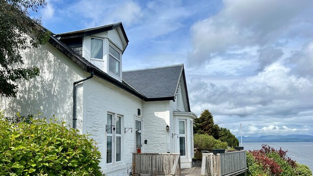 Een wit huis met een houten terras en uitzicht op zee, omringd door weelderige begroeiing.