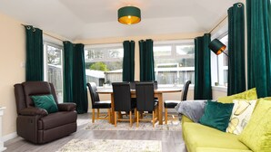 Un salon comprenant un fauteuil inclinable en cuir marron, un canapé jaune, une table à manger avec des chaises et une grande fenêtre ornée de rideaux verts.