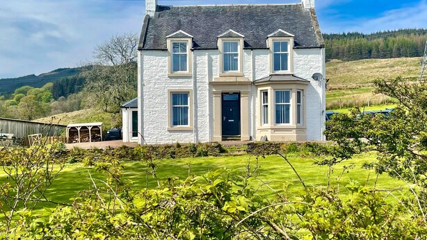 Une maison à deux étages, avec une façade blanche, de nombreuses fenêtres et un toit foncé.