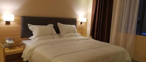 A hotel room with a large bed, two bedside lamps, a bedside phone, and a window with curtains.