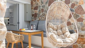 A modern kitchen with white cabinets, a stone wall, a wicker chair, and a dining table with fruit and a bottle. - Orama Milos (Milos)