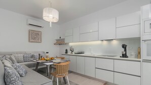A modern kitchen with white cabinets, a dining area with a sofa and table, and a hanging light fixture. - Orama Milos (Milos)