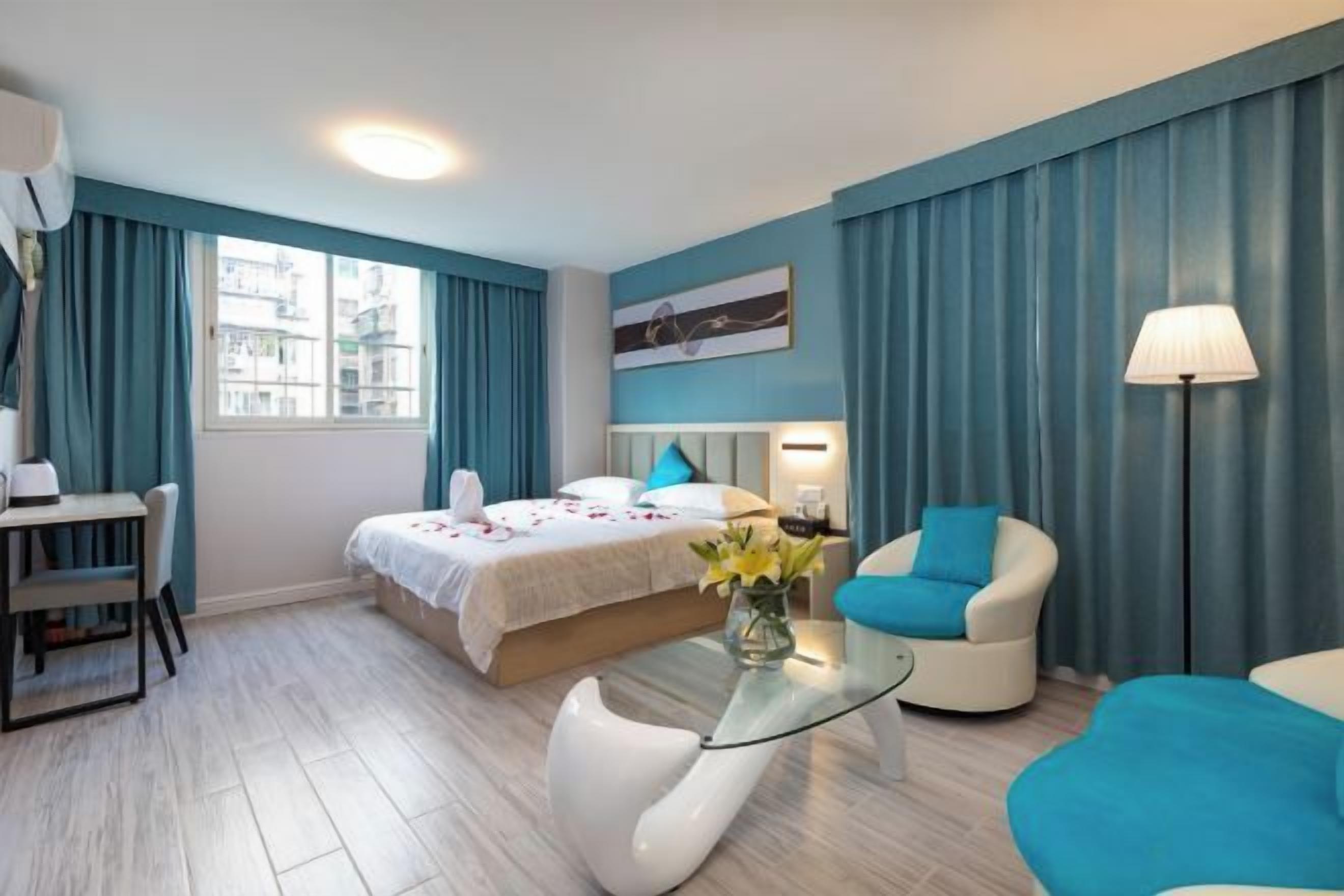 A hotel room with a bed, a desk, a chair, a glass coffee table, and a lamp.