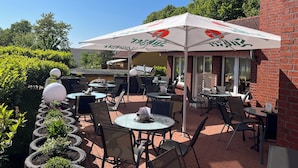 An outdoor patio with round tables and chairs, some under umbrellas, and a brick building in the background. - Hotel Nordwind Lohme (Lohme)
