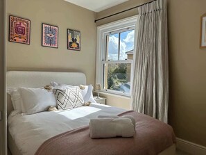 A bedroom with a bed, pillows, a bedside table, a window with curtains, and wall art.