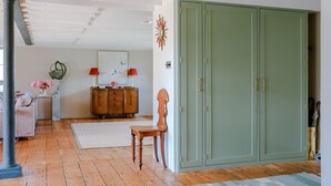 Una habitación con suelo de madera, un armario verde, una silla de madera, un tapete blanco, una lámpara roja y un reloj de pared.