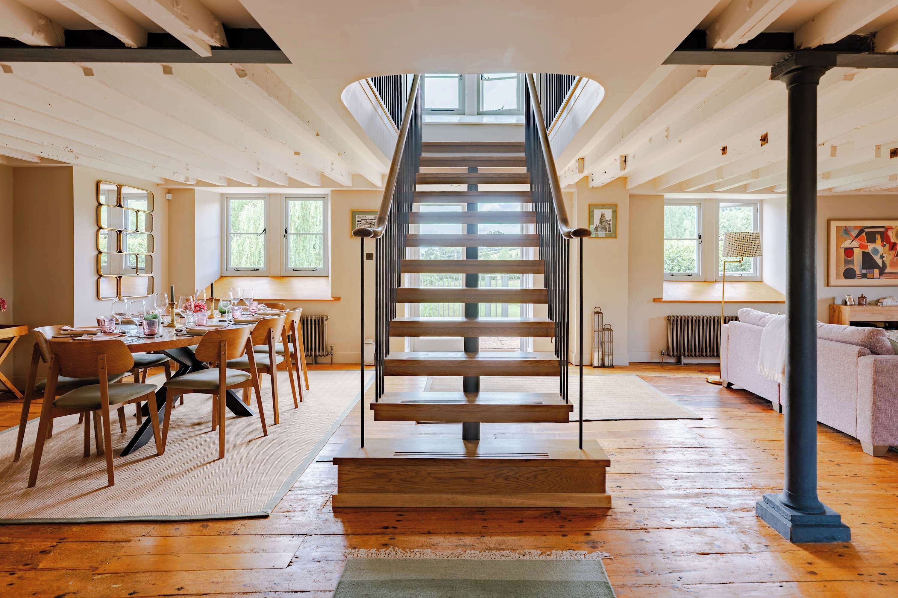 A staircase leading to an upper floor, wooden flooring, a dining area with chairs and a table, and a sofa in the living area.