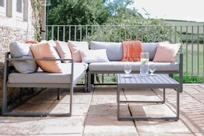 A modern outdoor seating area with a sofa and two tables, a metal railing, and a view of greenery. - Iron Mill House - Riverside Hot Tub - Frome (Somerset)