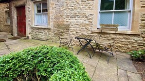 Une petite maison en pierre avec une porte rouge, deux fenêtres et un petit jardin agrémenté d’un banc et d’une table.