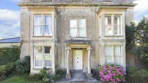 A two-story stone building with a central entrance flanked by columns, a symmetrical facade, and a small garden with pink flowers.