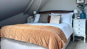 A bedroom with a sloped ceiling, a bed with a brown quilt, white bedding, a bedside table with a lamp, a small nightstand, and a framed picture on the wall. - Edwardian Town House - Garden & Cinema (Somerset)