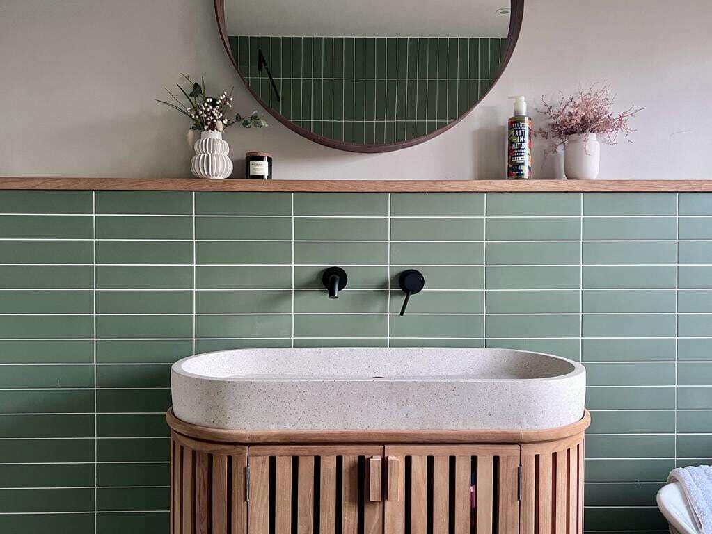 Een badkamer met een groen betegelde wand, een ronde spiegel, een houten wastafel en twee zwarte kraanwerk.