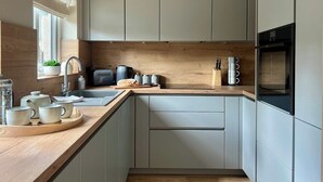 A modern kitchen with light grey cabinets, a wooden countertop, a sink, and a built-in oven. - Leaze Garden Cottage - Frome (Somerset)