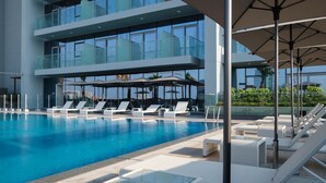 Ein moderner Hotelpoolbereich mit klarem, blauem Wasser, weißen Liegestühlen und einem Gebäude mit Balkonen, das von Glas umschlossen ist.