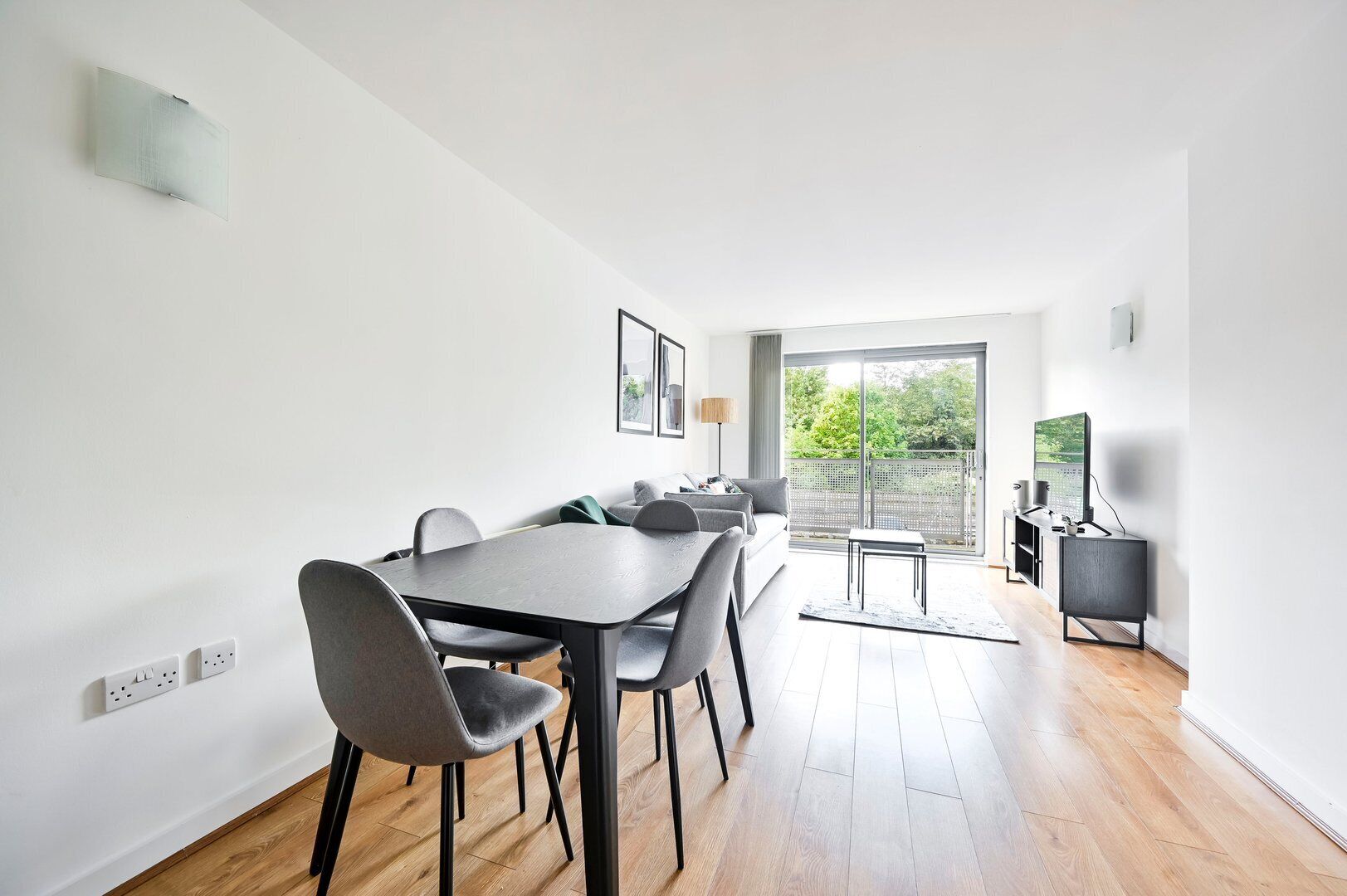 A modern dining area with a long table and chairs, a small seating area with a sofa and a lamp, and a view of greenery through a glass door.