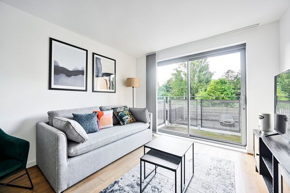 A modern living room with a grey sofa, a coffee table, and a large sliding glass door leading to a balcony. - GuestReady - Delightful retreat in Greenwich (London)