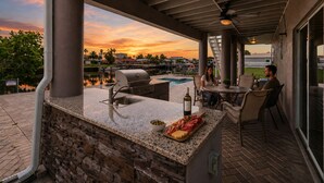 Een terras buiten met een barbecue, zitplaatsen en uitzicht op de zonsondergang boven het water.