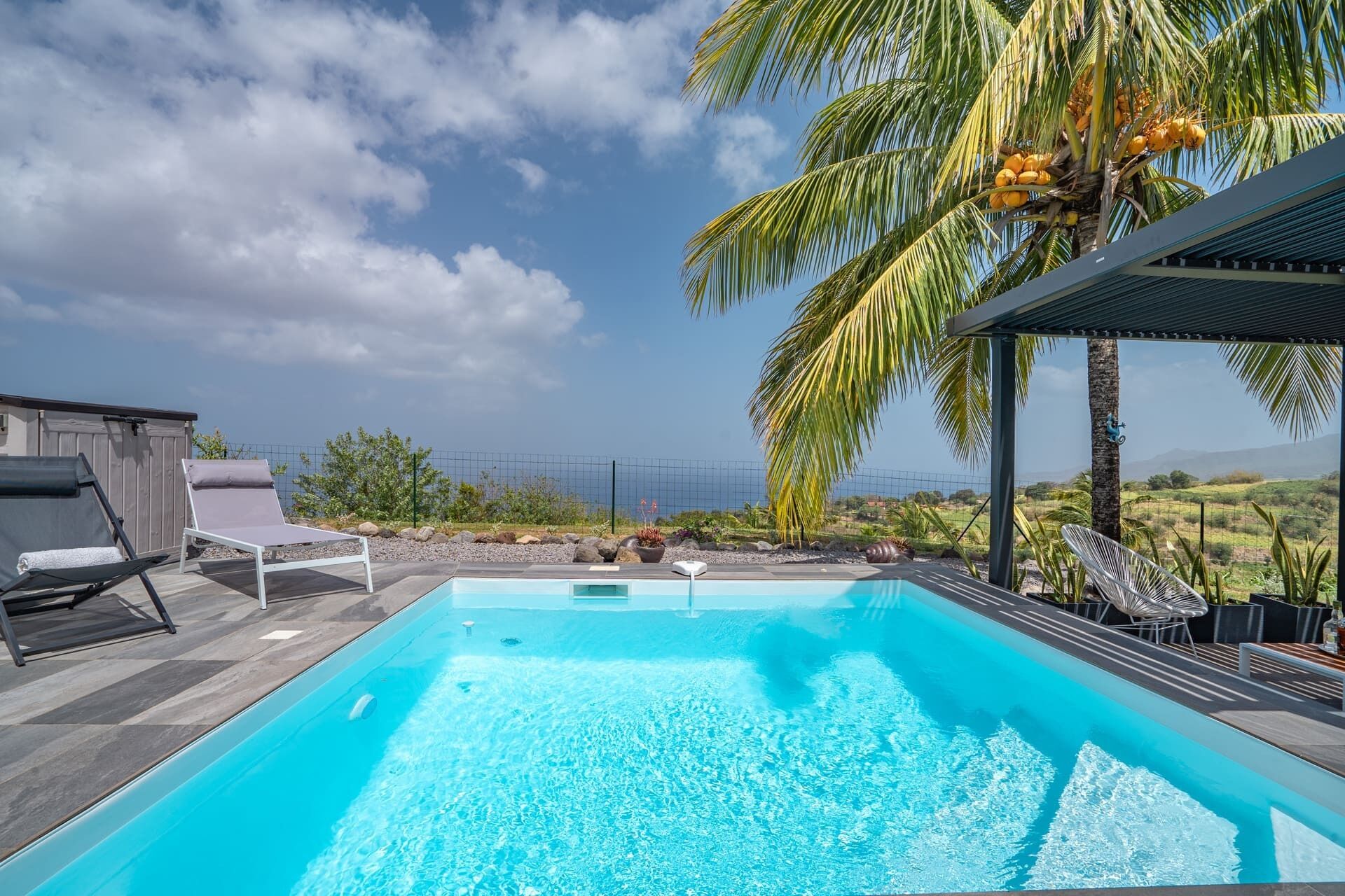 A rectangular swimming pool with clear blue water, surrounded by a wooden deck. There are lounge chairs and a covered seating area with a view of the landscape.