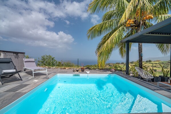 Una piscina rectangular con agua azul cristalina, rodeada por una terraza de madera. Hay sillones de descanso y un área de estar cubierta con vistas al paisaje.