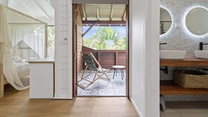 Una habitación con piso de madera, una cama con cabecera blanca, una mesita y una silla. Se ve un acceso a un área exterior con terraza de madera y vista a follaje.