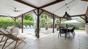Un espacio al aire libre techado con piscina, sillas de descanso, una mesa de comedor y ventiladores de techo.