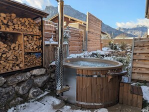 A wooden cabin with a hot tub, stacked firewood, and a stone wall. - "Heuberg" Apartment in the Inn Valley with a spa—central & quiet (Flintsbach)