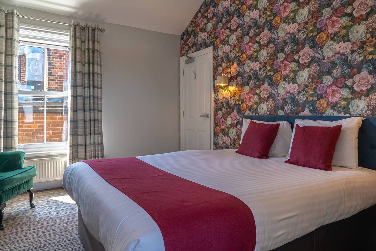 Une chambre à coucher avec un grand lit, un fauteuil vert, un papier peint à motifs floraux et une fenêtre avec des rideaux.