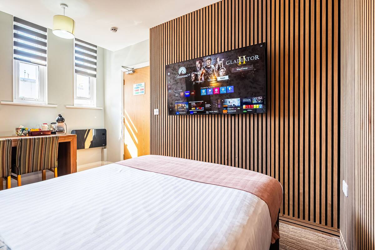 Une chambre d’hôtel dotée d’un grand téléviseur à écran plat fixé au mur, d’un lit avec une couvre-lit à rayures, d’un bureau en bois avec une chaise et d’une fenêtre avec des stores.