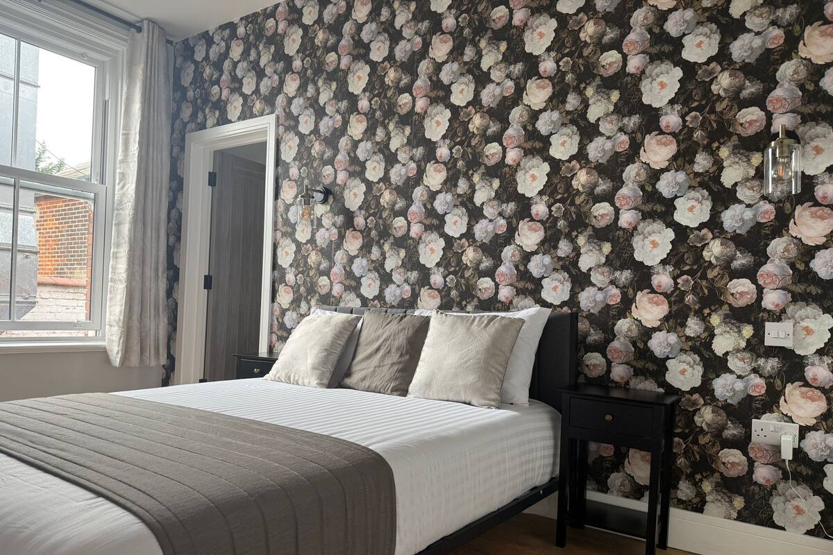 Une chambre à coucher avec un papier peint à motifs floraux, un lit recouvert d’une couette grise et blanche, une table de chevet et une fenêtre avec des rideaux.